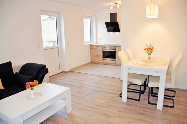 A modern living room with a white sofa, a dining table with chairs, and a kitchen area in the background.