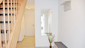 Een gang met een houten trap, een bankje, een spiegel en een wandplank met haken.