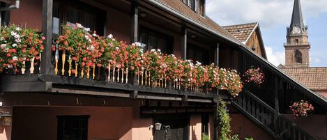 A traditional building with a balcony adorned with flowers, a visible staircase, and a church steeple in the background.