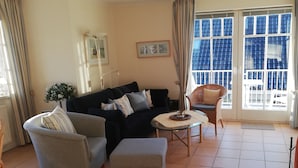 A living room with a sofa, armchair, ottoman, and a small table. There is a window with blue shutters and a door with curtains. - Johannesstr. 12 apt. 4 (Wyk auf Föhr)