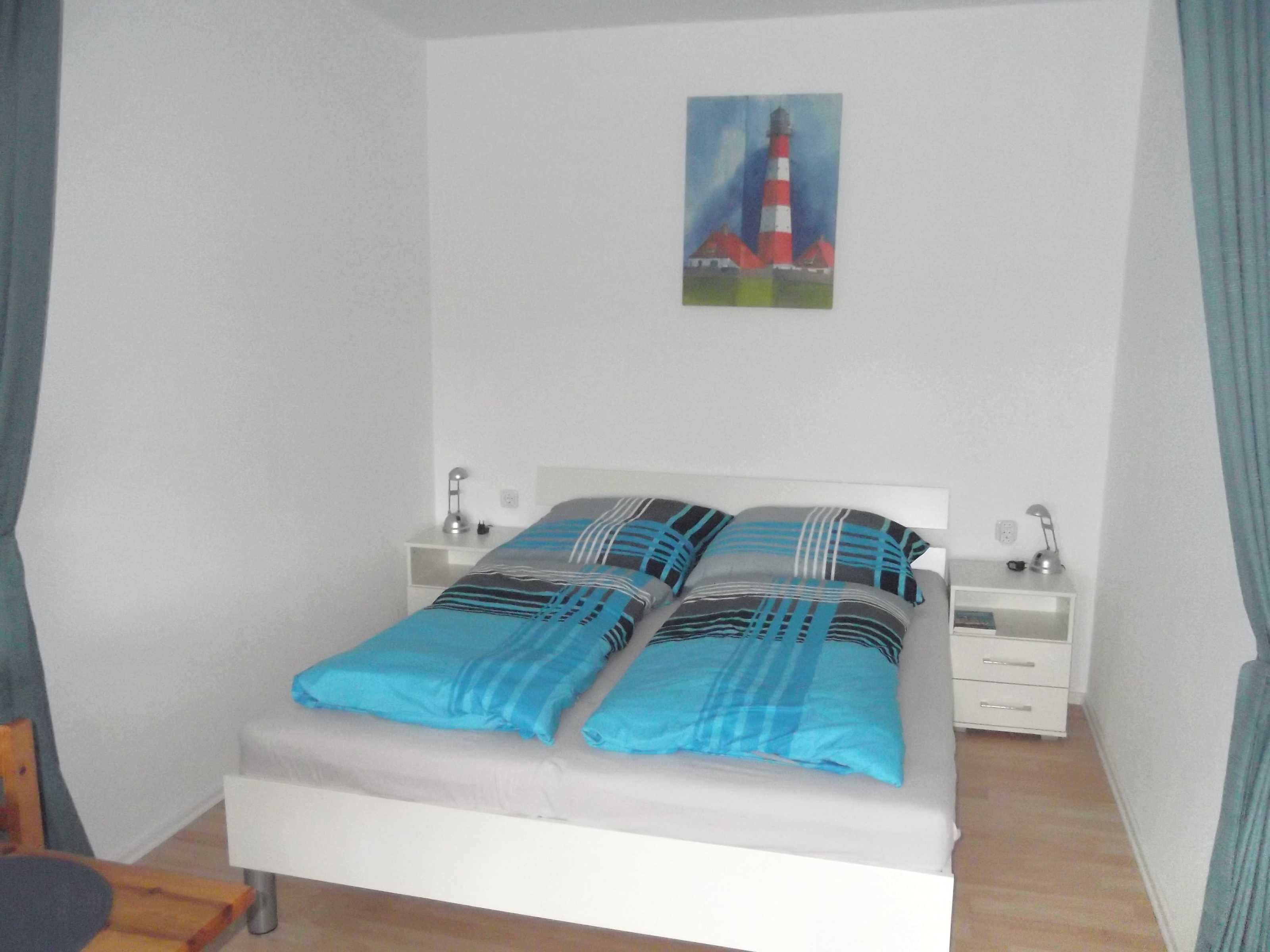 Ein ordentlich gemachtes Bett mit blau-schwarz gestreifter Bettwäsche, zwei Nachttische mit Lampen und ein Gemälde eines Leuchtturms an der Wand.
