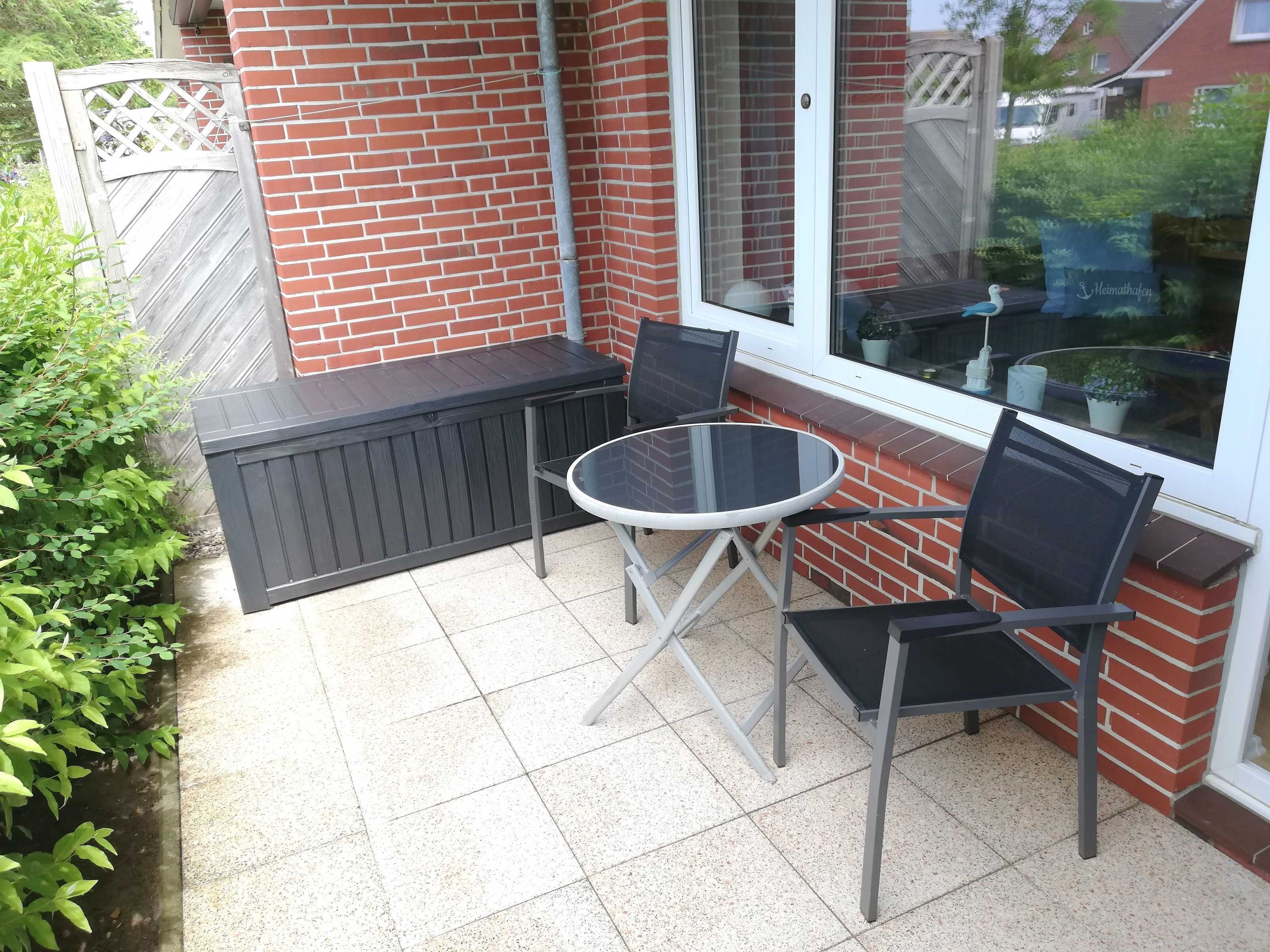 Eine Terrasse mit einem runden Glastisch und zwei Stühlen, einer Ziegelwand und einem Fenster mit Blick auf die Nachbarschaft.