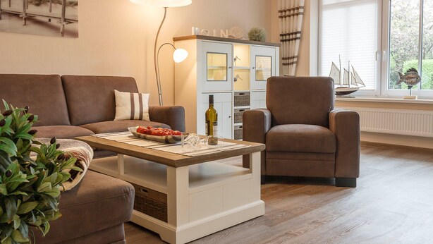 A living room with a brown sofa, a wooden coffee table with a bottle and fruit, a white cabinet with glass doors, and a window with a view of greenery. - Apple blossom house, Schmalstieg 1 (Wyk auf Föhr)