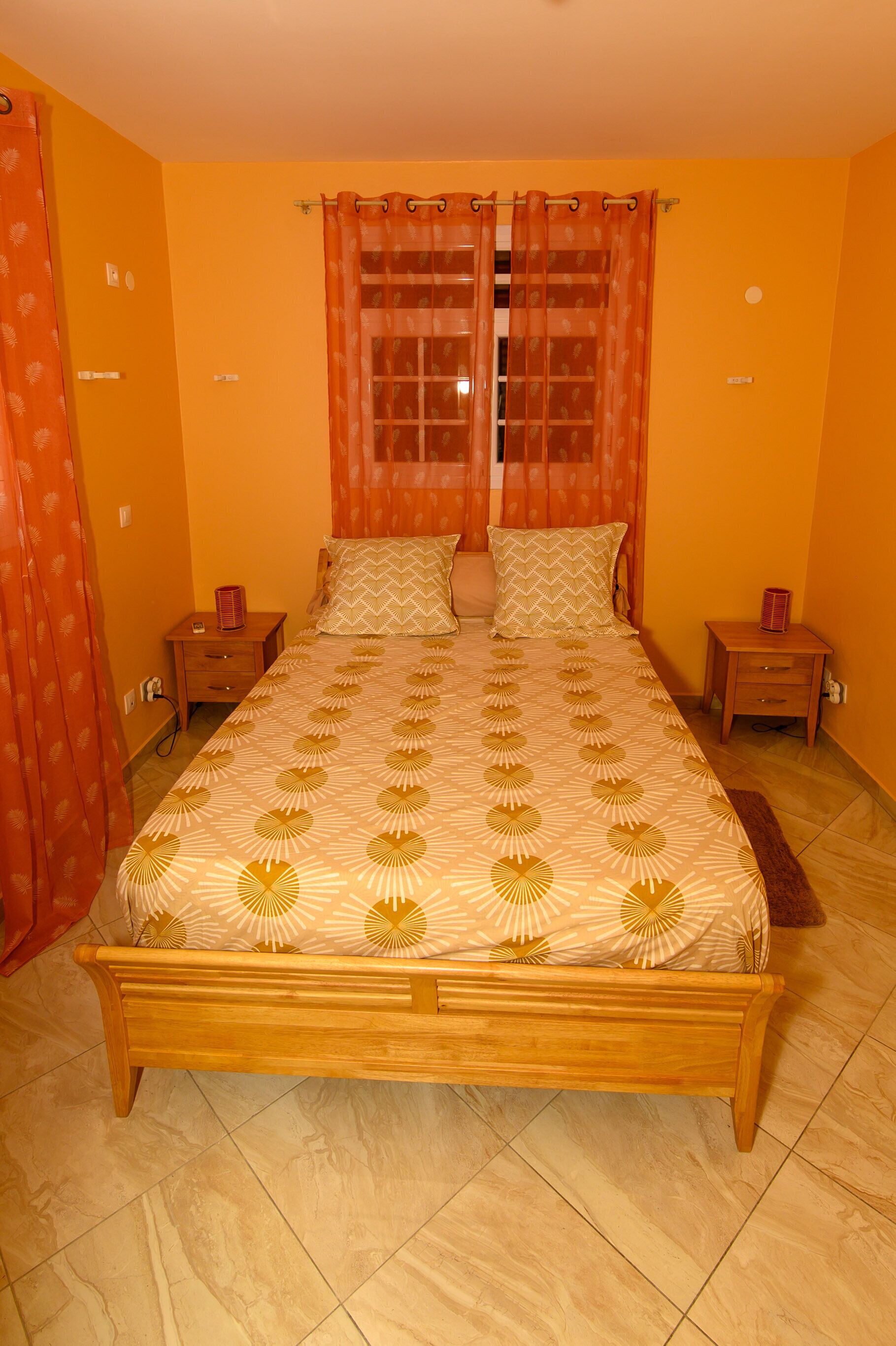 Un dormitorio con cama de madera, cortinas naranjas y suelo de baldosas.