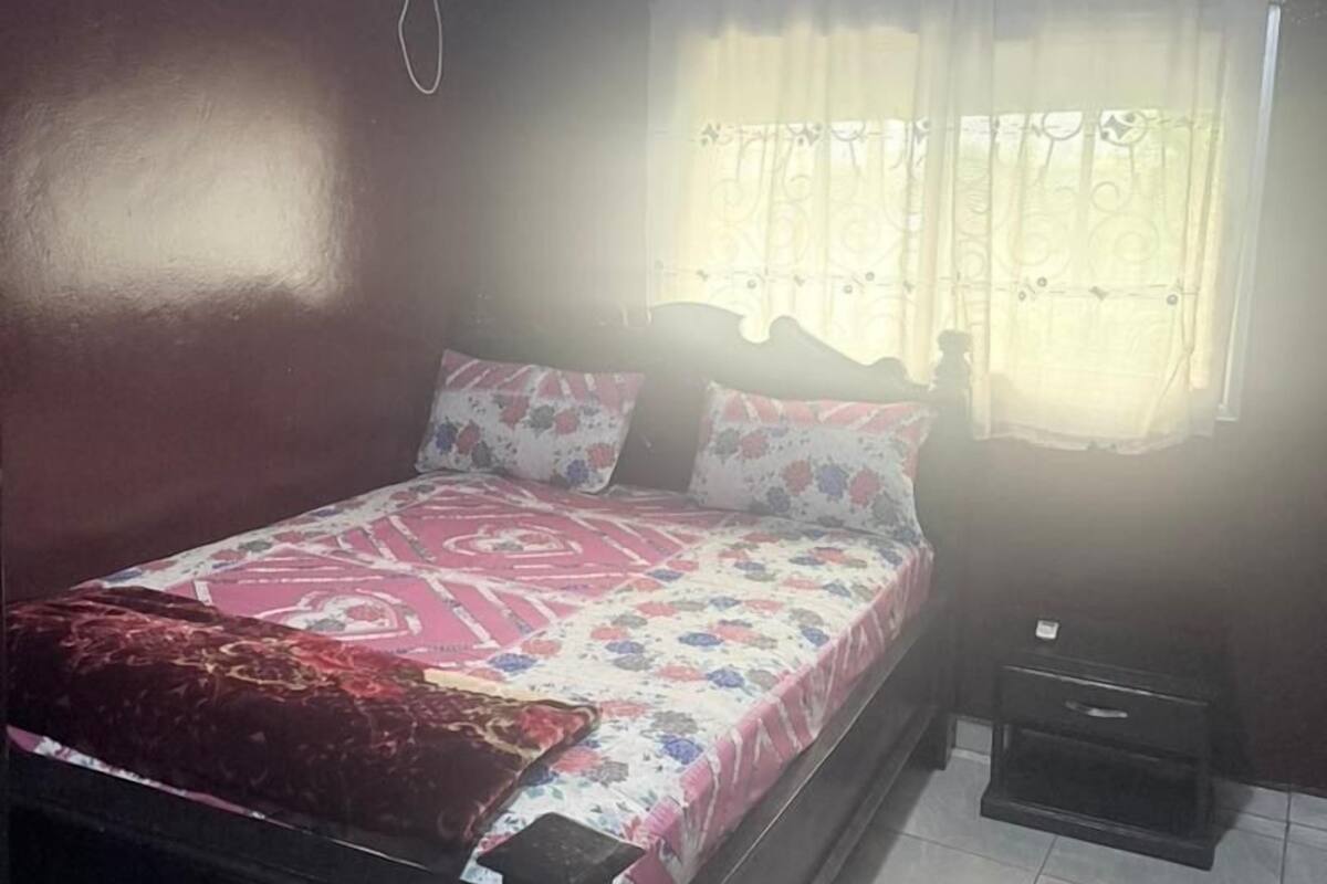 Quarto com cama, ar condicionado, janela com cortinas e mesa de cabeceira.