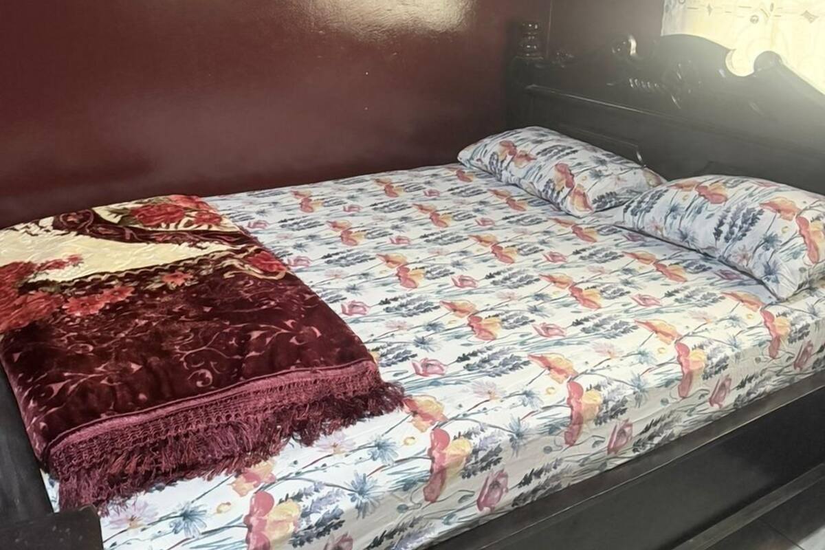 Uma cama de solteiro com colcha floral e manta vermelha, um aparelho de ar condicionado branco na parede, e uma janela com cortina.