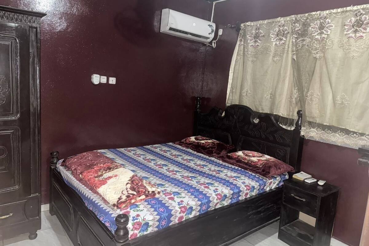 Quarto com cama de madeira escura, colcha estampada, mesa de cabeceira e ar-condicionado fixado na parede.