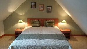 Quarto com teto inclinado, duas luminárias de cabeceira, cama com travesseiro listrado e três quadros na parede.