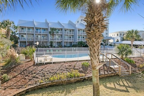 A hotel with a swimming pool, surrounded by a white fence, palm trees, and a well-maintained garden.