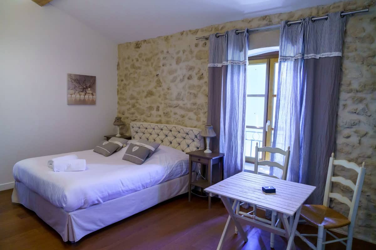 A bedroom with a bed, a table, and a chair. There is a window with curtains and a stone wall.