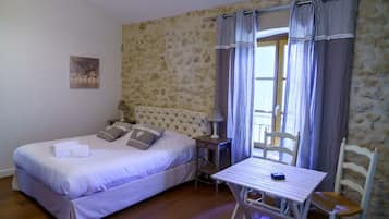 A bedroom with a bed, a table, and a chair. There is a window with curtains and a stone wall.