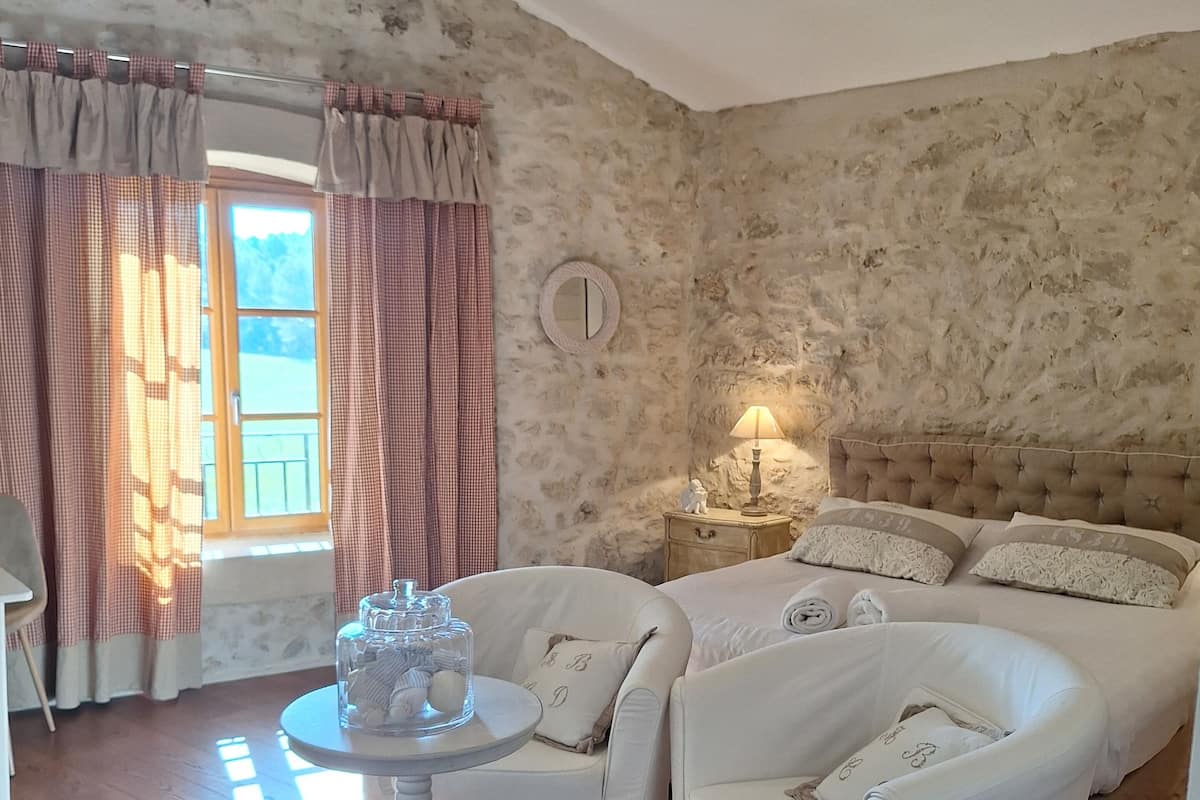 A room with a stone wall, a bed with a headboard, a bedside table with a lamp, and a chair.