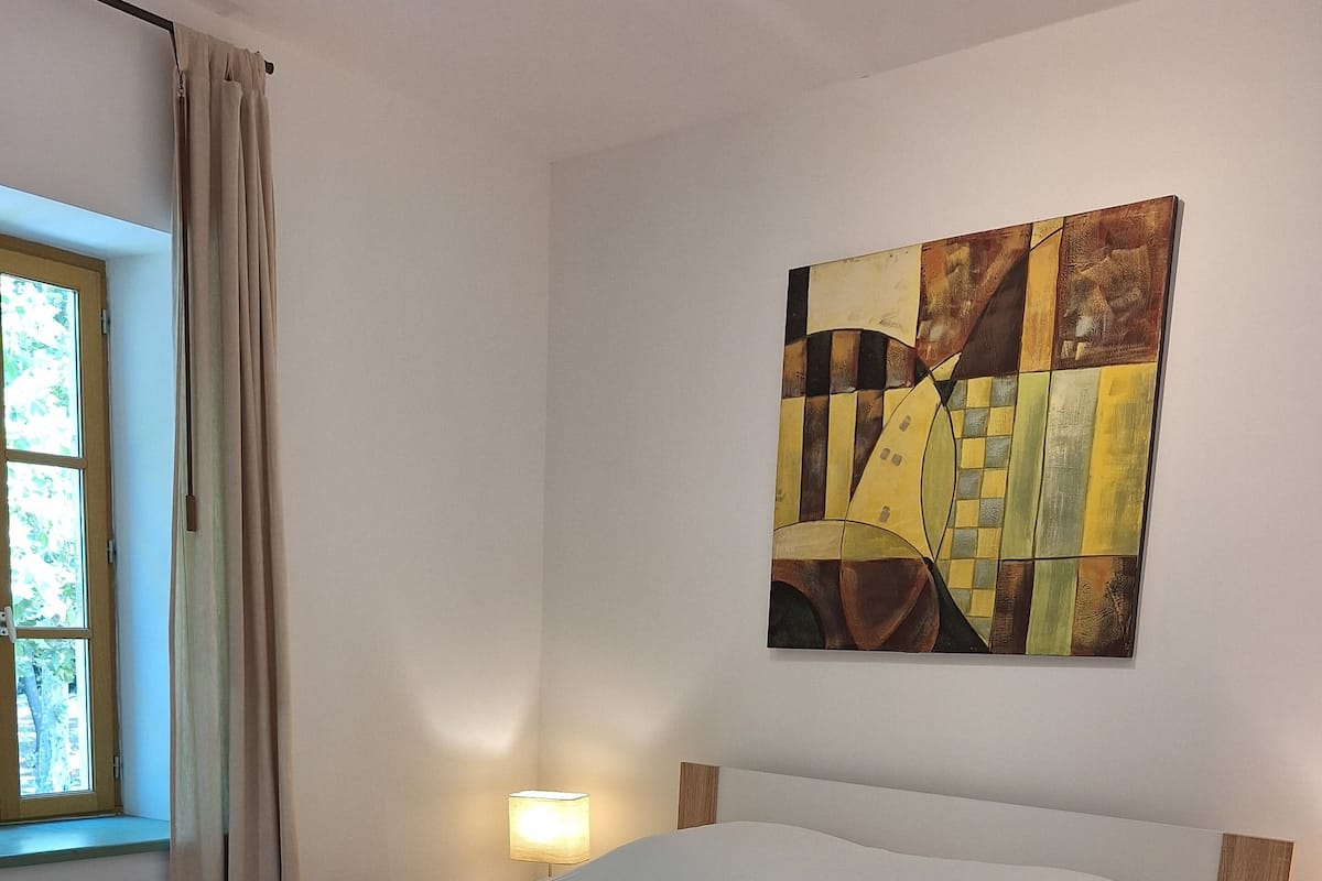 A neatly made bed with white linens and rolled towels, a bedside lamp, and a large abstract painting on the wall.