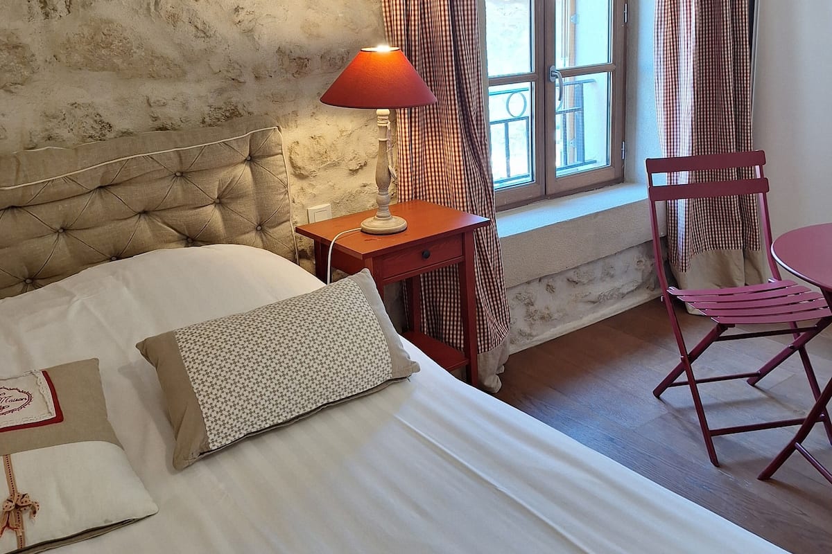 A bedroom with a bed, a nightstand, a lamp, a chair, and a window with curtains.