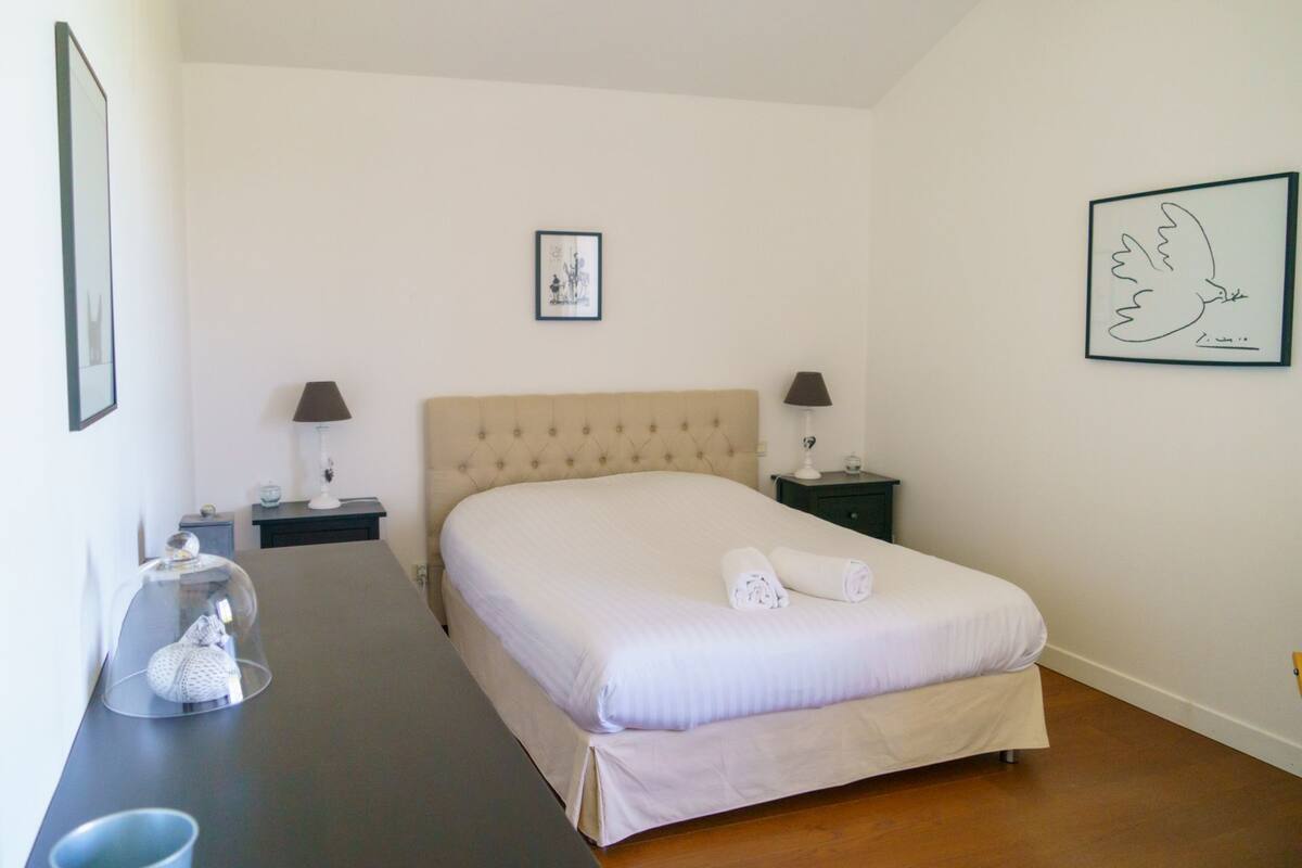 A neatly arranged bedroom with a large bed, two bedside tables with lamps, a framed picture on the wall, and a small table with a glass dome containing a decorative item.
