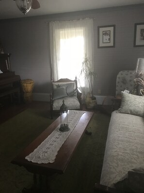 Een woonkamer met een piano, een bank, een tafel met een kantkleed en een lamp.