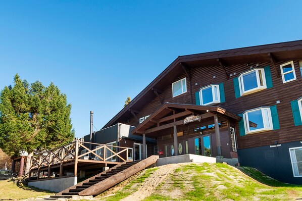 Un edificio de madera de varios pisos con una escalera que conduce a la entrada, rodeado de árboles y un área verde.