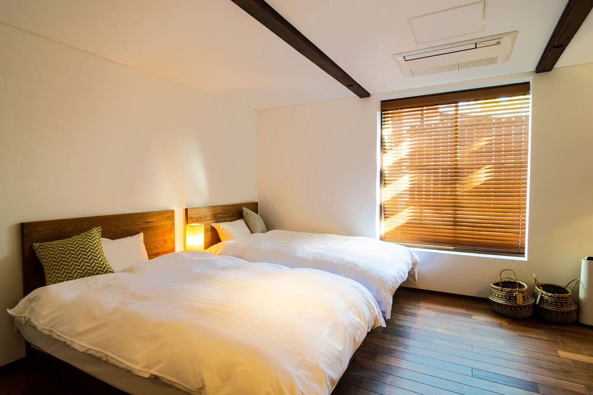 A bedroom with two beds, wooden headboards, a window with blinds, and a ceiling vent.