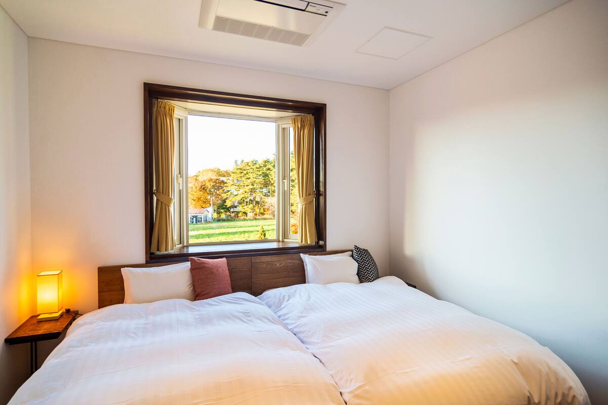 A hotel room with a large bed, a nightstand with a lamp, a window with curtains, and a view of trees outside.