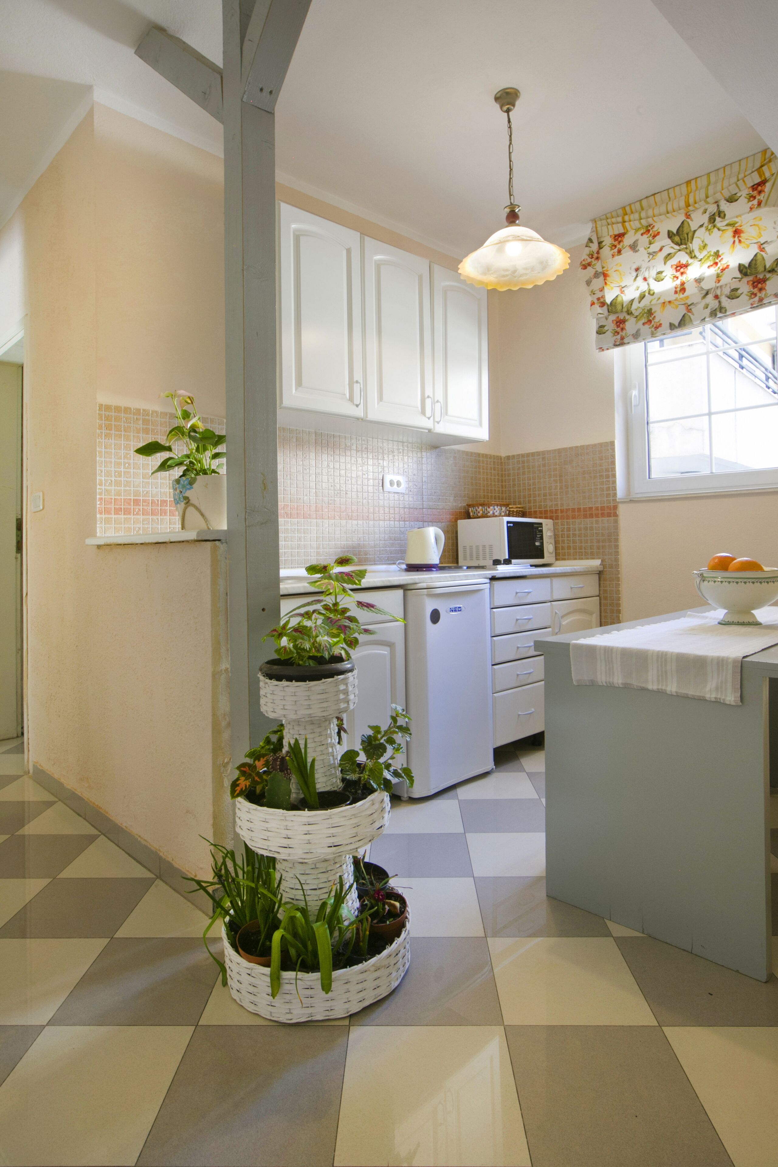 A kitchen with white cabinets, a microwave, a sink, and a window with floral curtains. There are potted plants on a stand and a checkered floor.