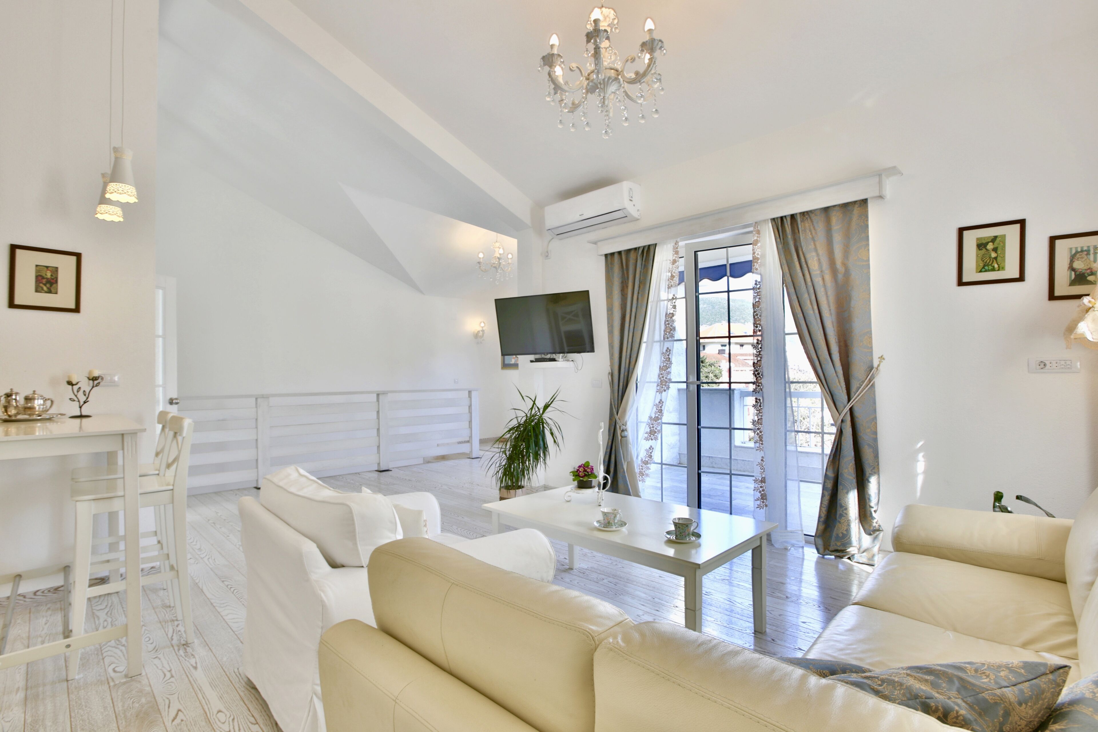 Uma sala de estar ampla com teto alto, um candelabro, uma TV de tela plana, um espaço de refeições com mesa e cadeiras, e uma janela grande com cortinas.