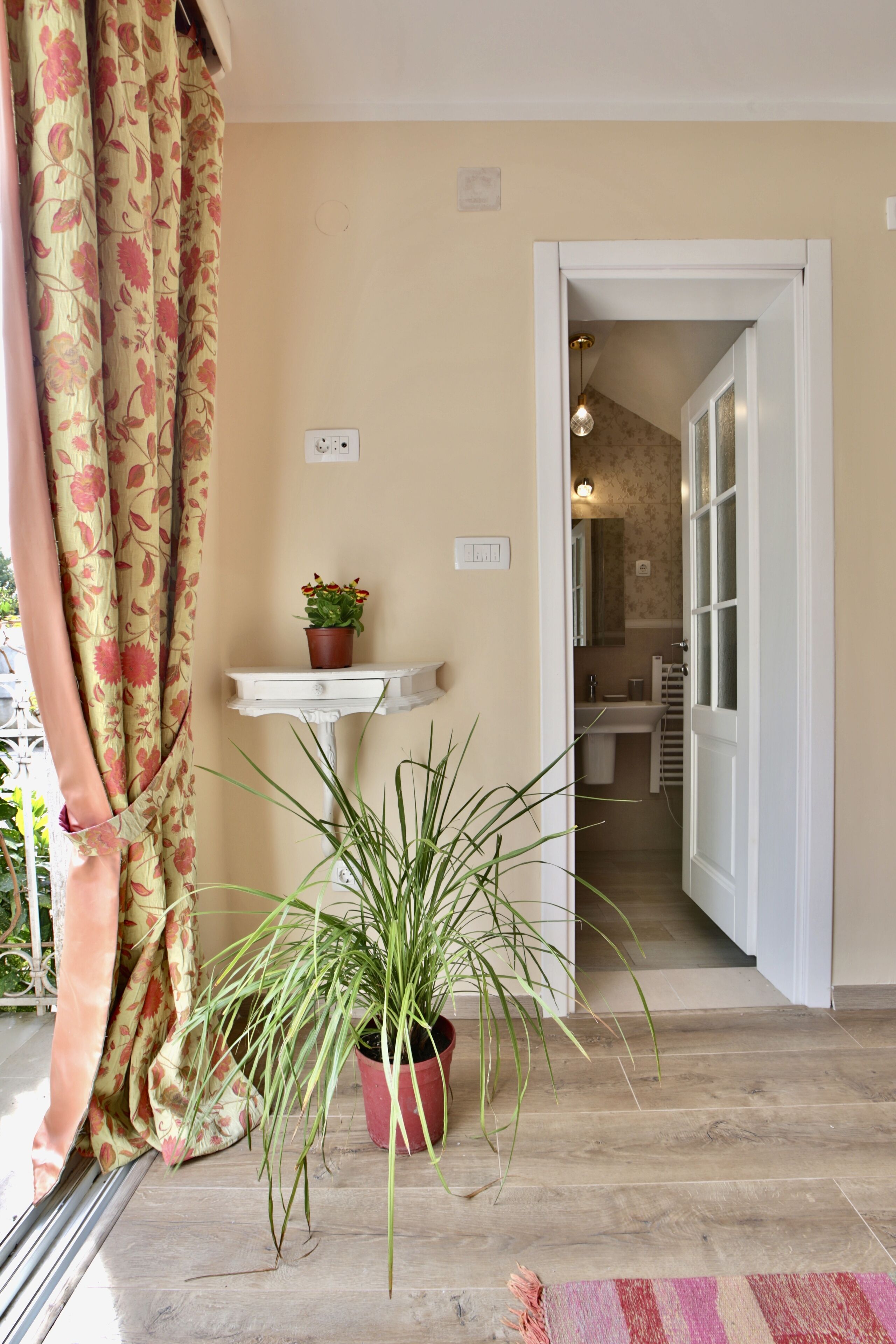 A room with a wooden floor, a potted plant, a small white pedestal table with a flower pot, and a doorway leading to a bathroom.