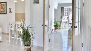 A spacious room with a wooden floor, a dining area with white chairs, a potted plant, and a large window with a view of buildings. - Marinero Apartments (Budva)
