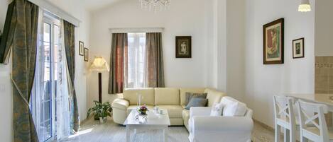 Un salon spacieux avec un canapé blanc, une table basse et un lustre. Les fenêtres sont dotées de rideaux, il y a une lampe sur pied et des tableaux encadrés ornent les murs.