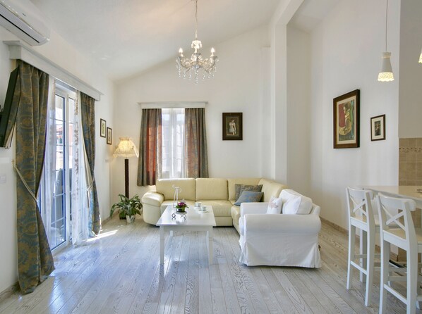 Amplia sala de estar con un sofá blanco, una mesa de centro y una araña. Hay cortinas en las ventanas, una lámpara de pie e imágenes enmarcadas en las paredes.