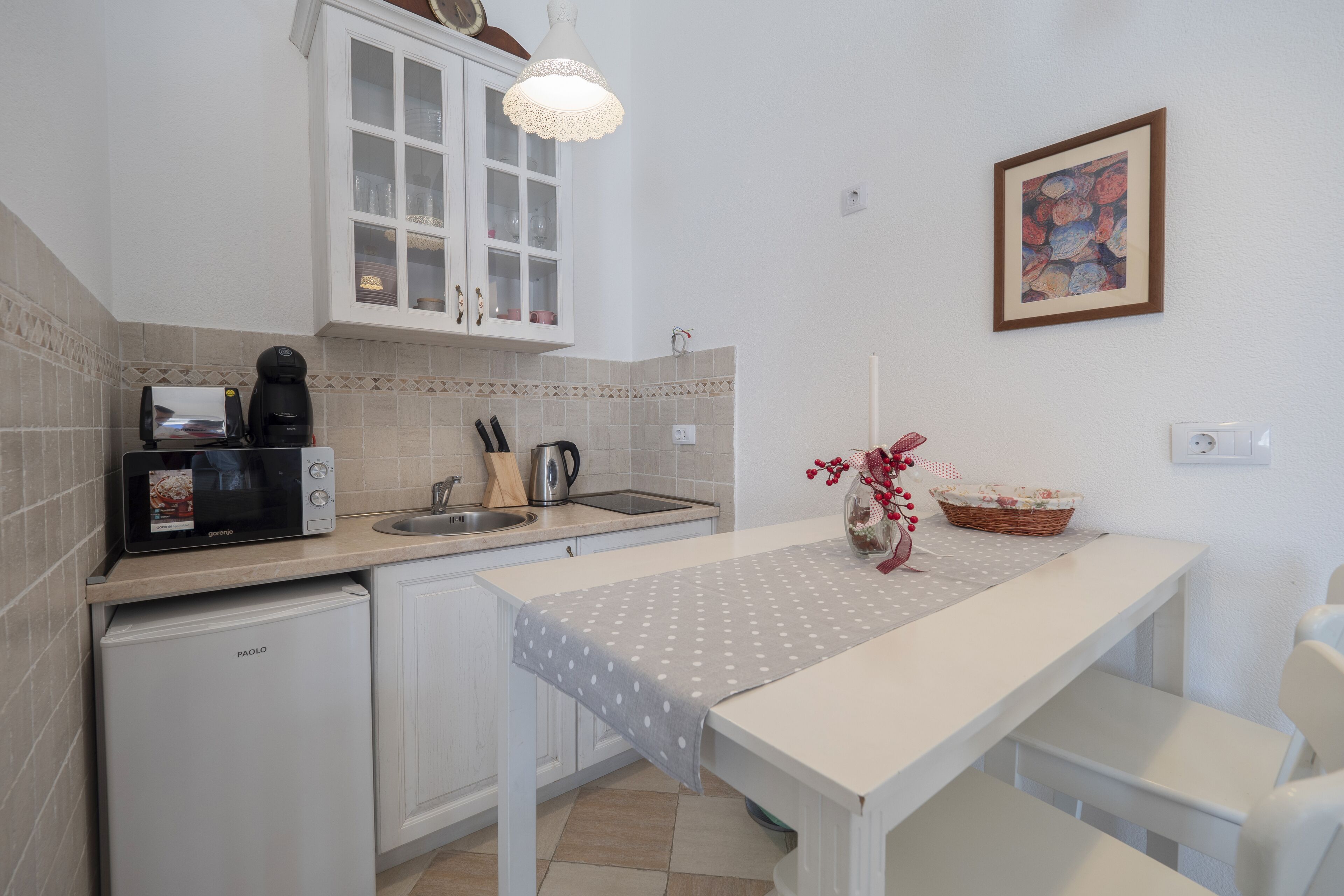 Een kleine keuken met een magnetron, koffiezetapparaat en spoelbak. Er is een kleine koelkast, een tafel met een tafelkleed met stipjes en een vaas met bloemen.