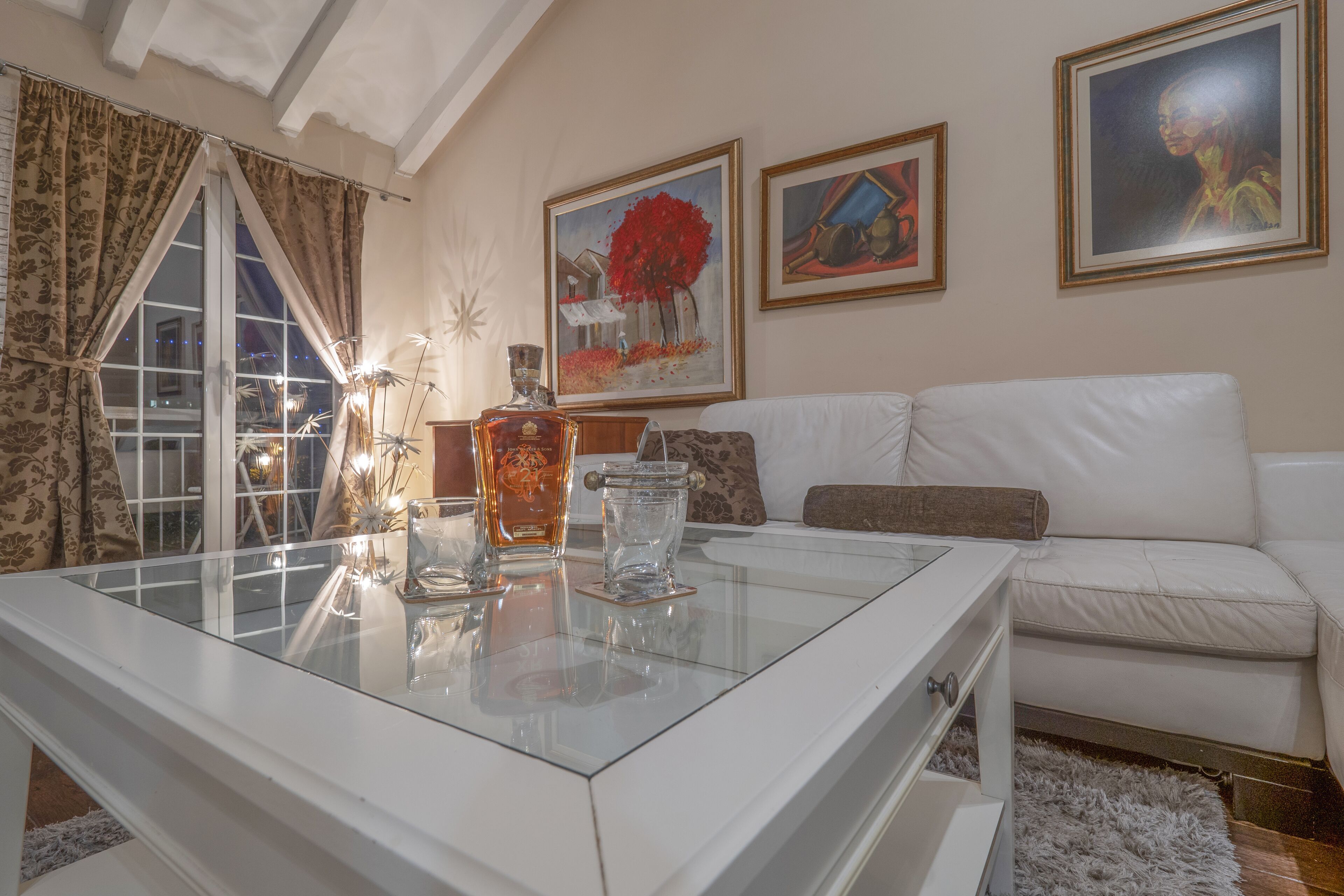 A modern living room with a glass coffee table, white sofa, and artwork on the walls.