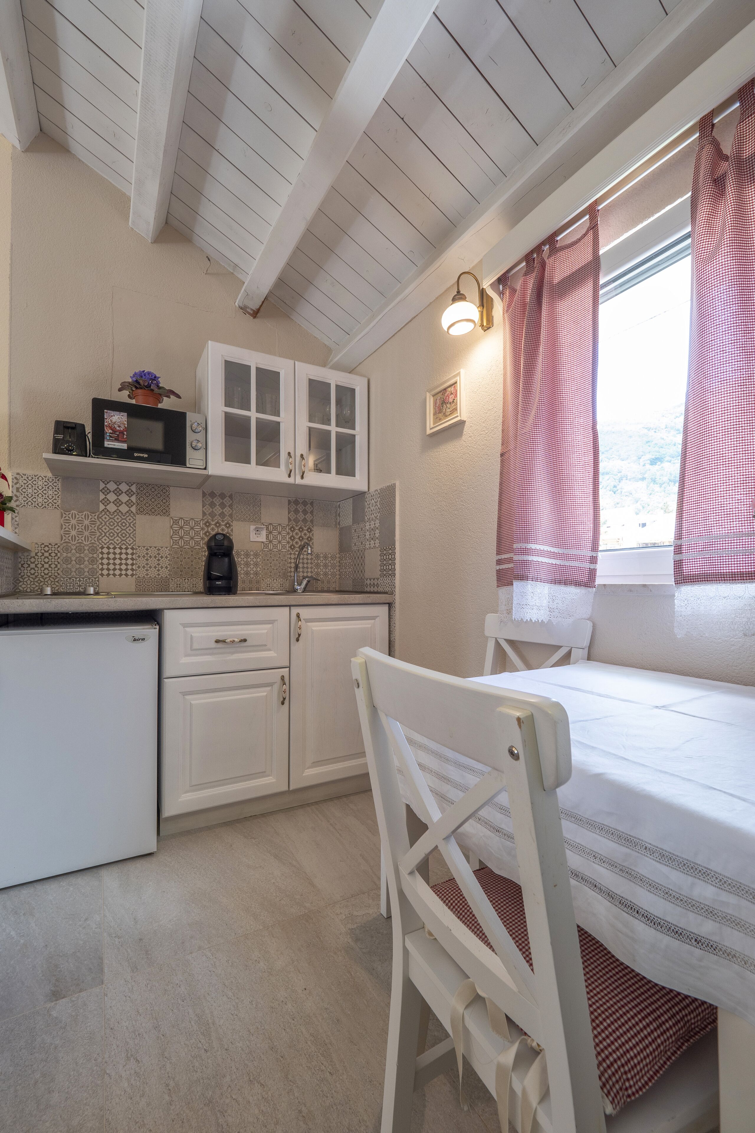 A compact kitchen with white cabinets, a microwave, and a coffee maker. There is a dining area with a white table and chair, and a window with red curtains.