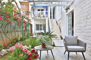 Un espacio al aire libre con una silla de mimbre y una mesa, rodeado de macetas con plantas y flores.