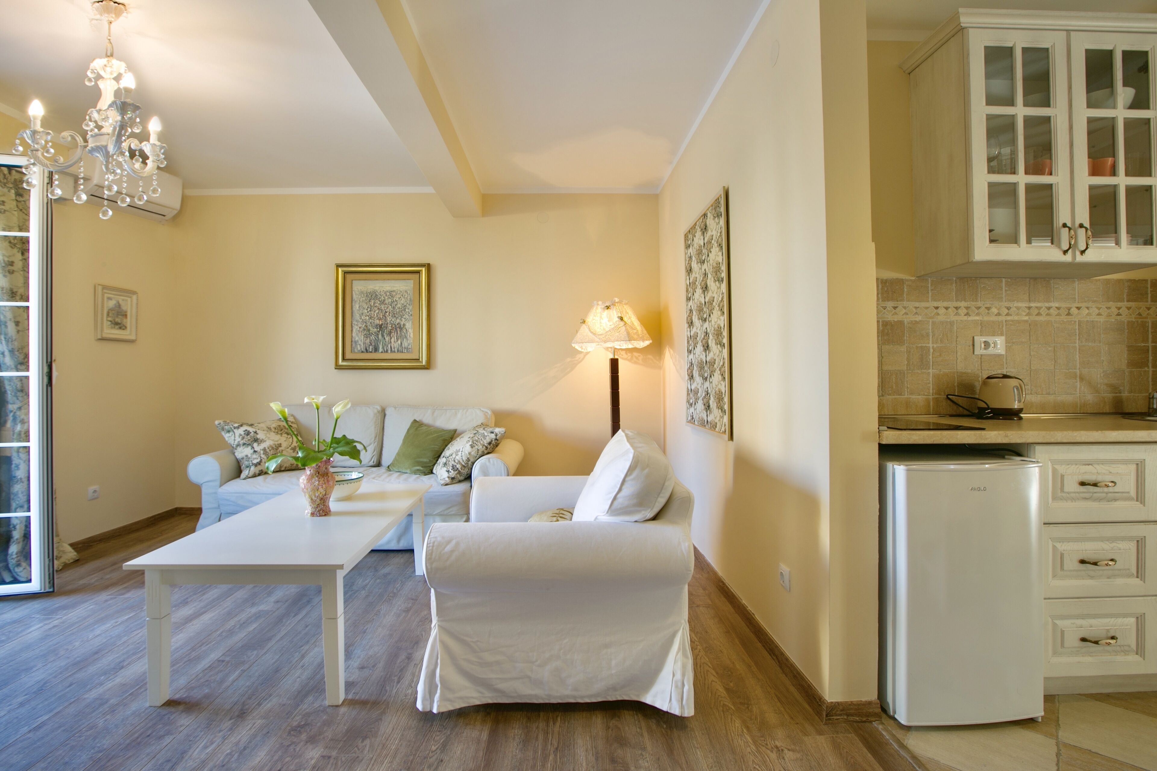 A living room with a sofa, a coffee table, and a lamp. There is a kitchen area with cabinets and a refrigerator.