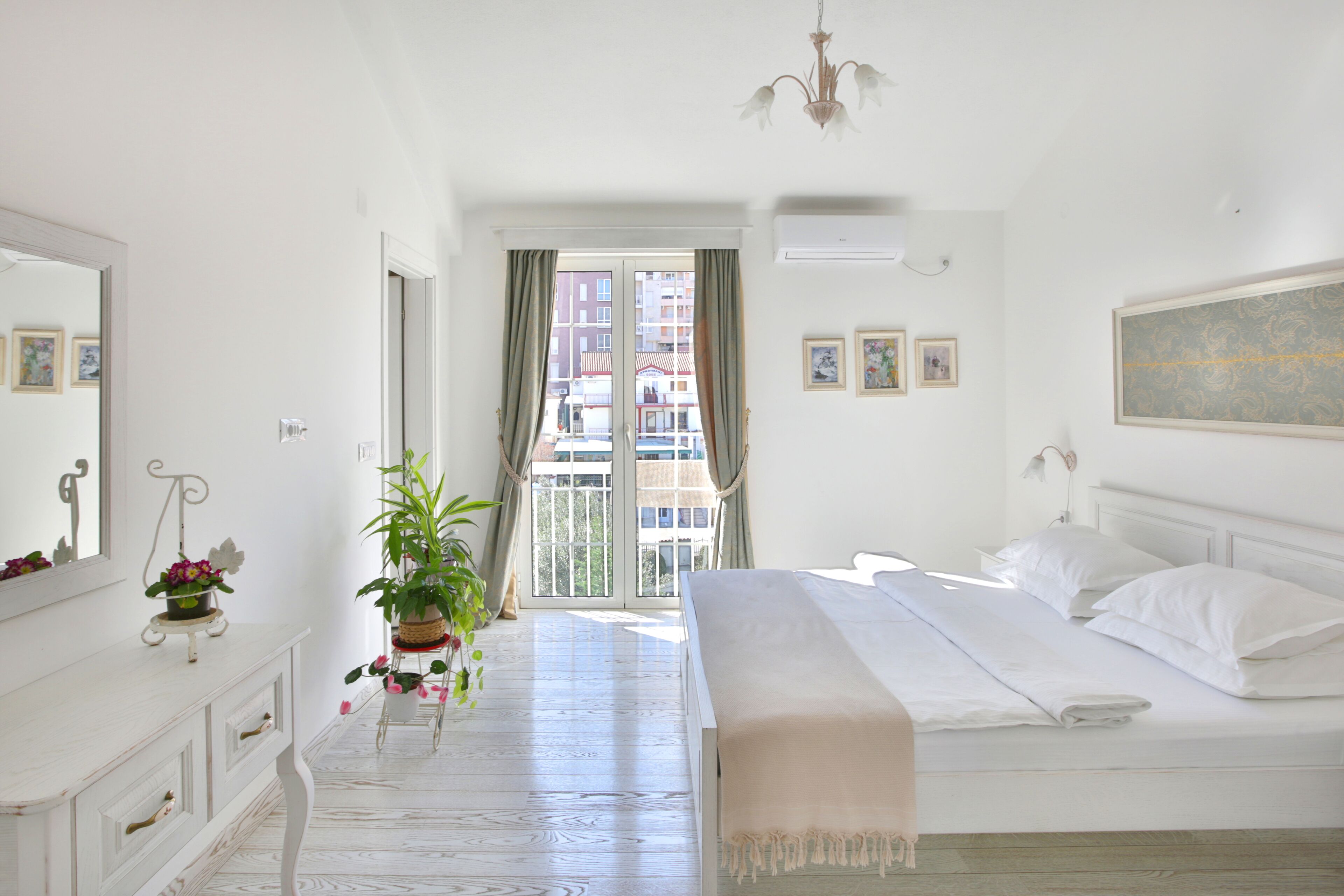 A bedroom with a bed, a dresser, a mirror, a potted plant, and a view of buildings through a balcony door.