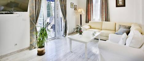 A modern living room with a white sofa, a coffee table, and a TV. There is a potted plant and a chandelier.