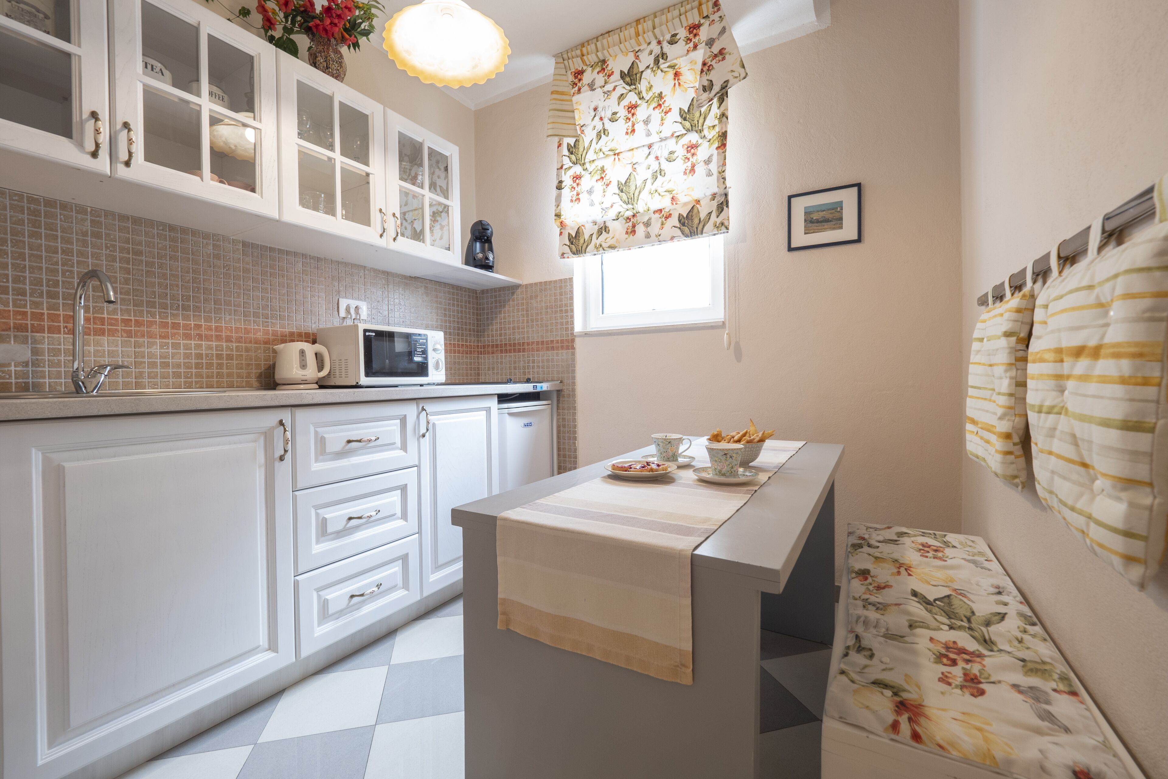 A kitchen with white cabinets, a microwave, a kettle, and a window with floral curtains.
