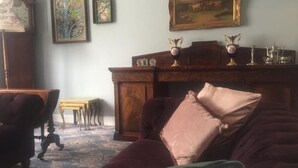 Una habitación con un sofá rojo oscuro, un sillón verde, una mesa de centro de madera, una chimenea con adornos y obras de arte enmarcadas en la pared.