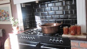 Una cocina con salpicadero de azulejos negros, una estufa y un horno de ladrillo. Hay frascos de conservas en la encimera y una olla de cobre sobre la estufa.