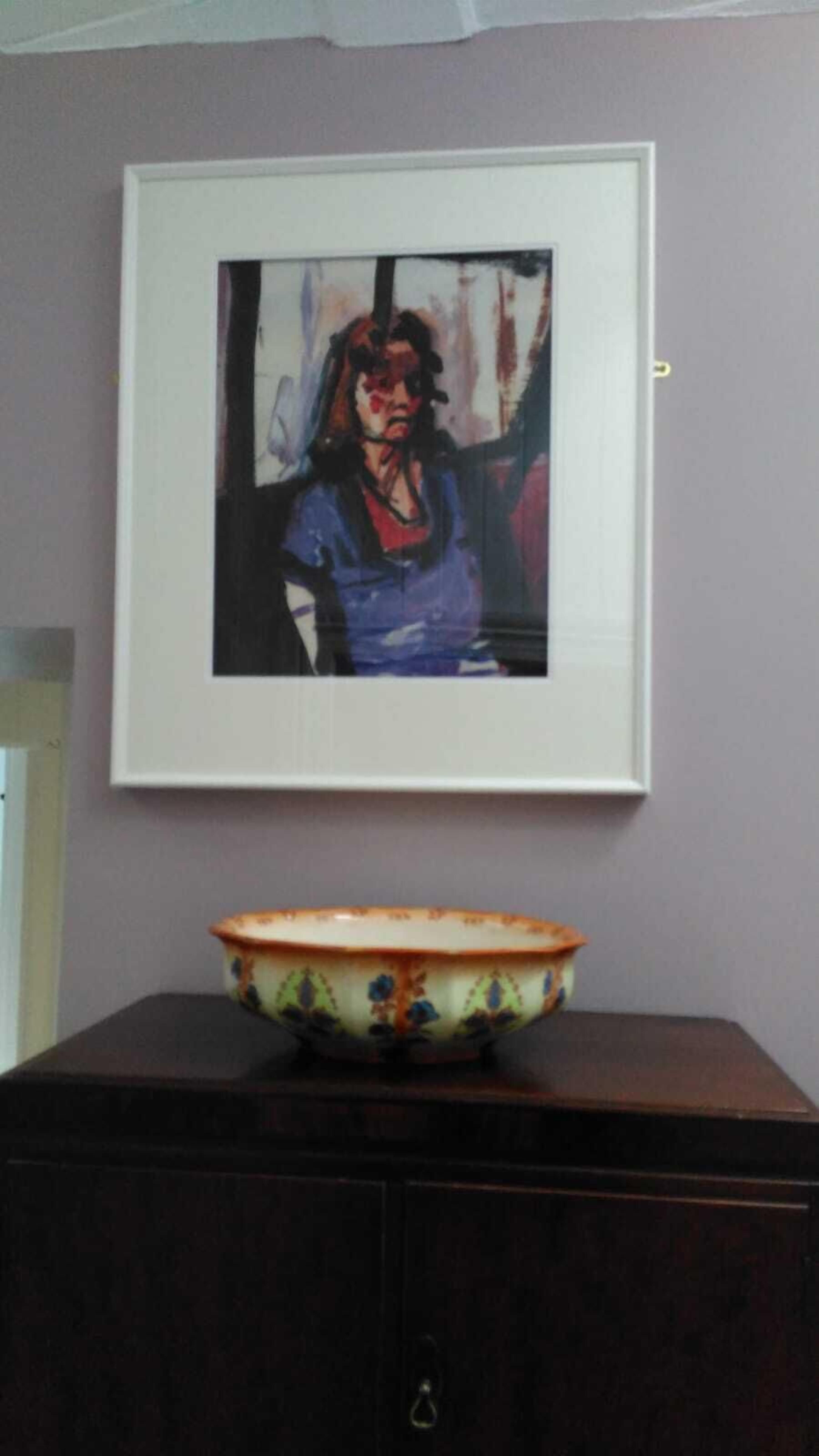 Una obra de arte en un marco sobre una pared, encima de un aparador de madera oscura con un cuenco decorativo.