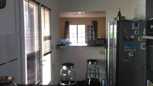 A compact kitchen with a refrigerator, two stools, a window with blinds, and a small shelf with a bowl. - The waterfall balcony (Trans-en-Provence)