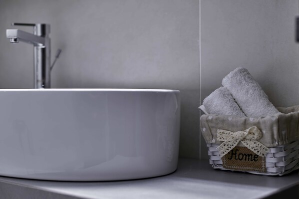 Un lavabo blanc dans la salle de bain, doté d’un robinet moderne, un panier en osier contenant des serviettes et une petite étiquette sur laquelle on peut lire « Chez soi ».