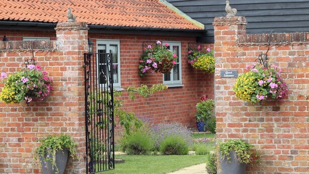 Una casa de ladrillo con techo de tejas, entrada con puerta y cestas de flores colgantes, además de plantas en macetas.