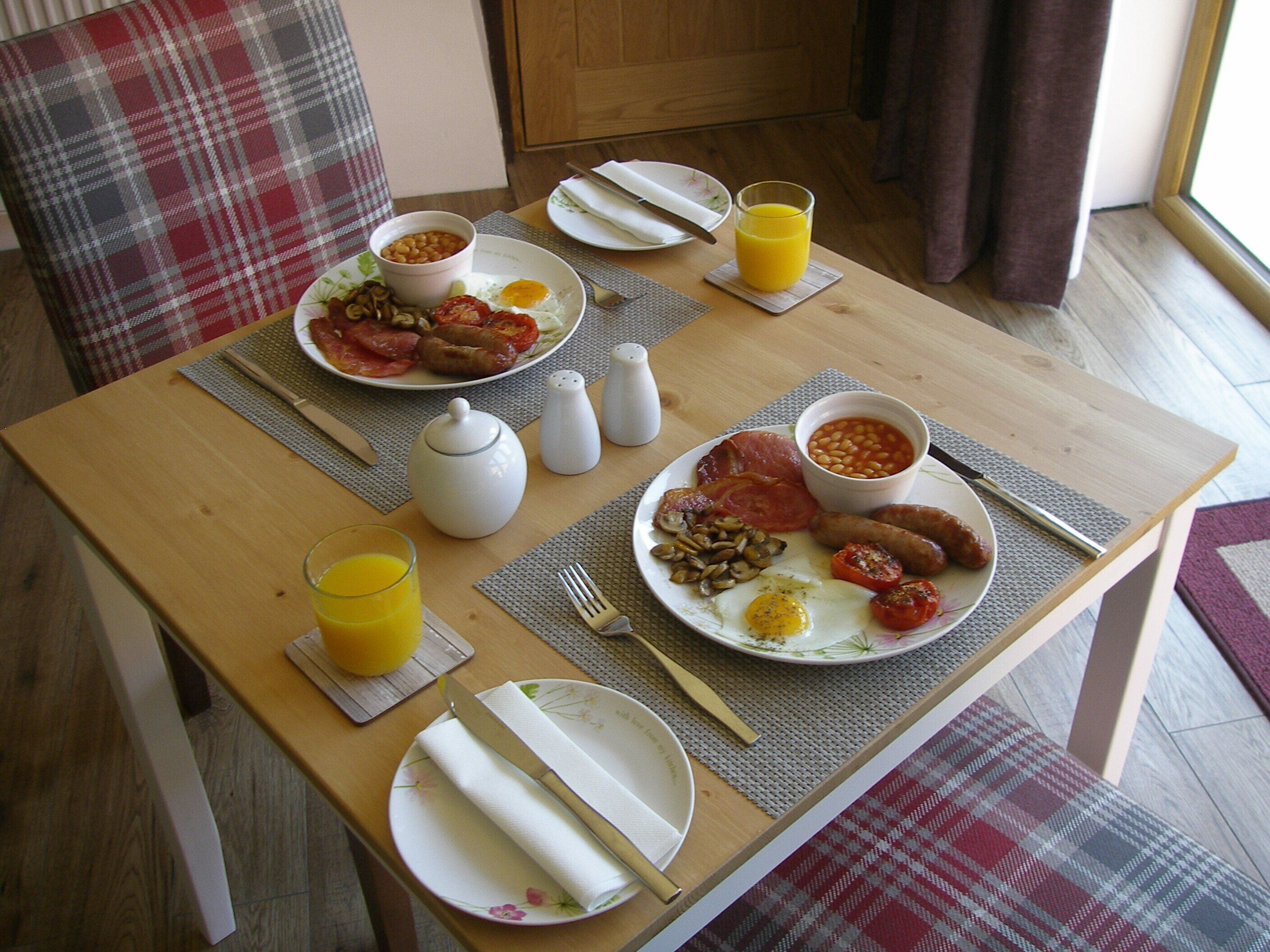 A breakfast table with plates of eggs, sausages, beans, and tomatoes, along with a glass of orange juice.
