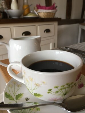 A cup of coffee on a saucer with floral designs, a spoon, and a white pitcher on a table. - Grove Barn Bed and Breakfast (Harleston)