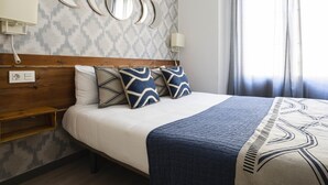A neatly made bed with a patterned bedspread and pillows, a wooden headboard, a wall-mounted mirror, and a window with curtains.