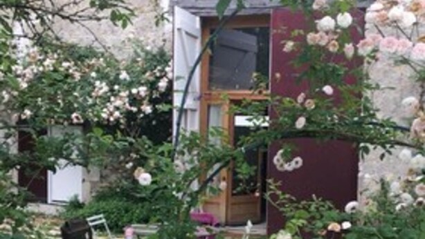 Um edifício rústico de pedra com porta e janela de madeira, rodeado por vegetação exuberante e flores em flor.