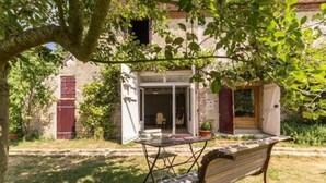 Uma casa rústica com jardim, um banco de madeira e uma mesa com cadeiras.
