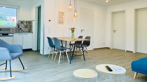 Eine moderne Küche mit weißen Schränken, ein Essbereich mit einem Holztisch und Stühlen, und ein Wohnbereich mit einem blauen Sofa und einem kleinen runden Tisch.