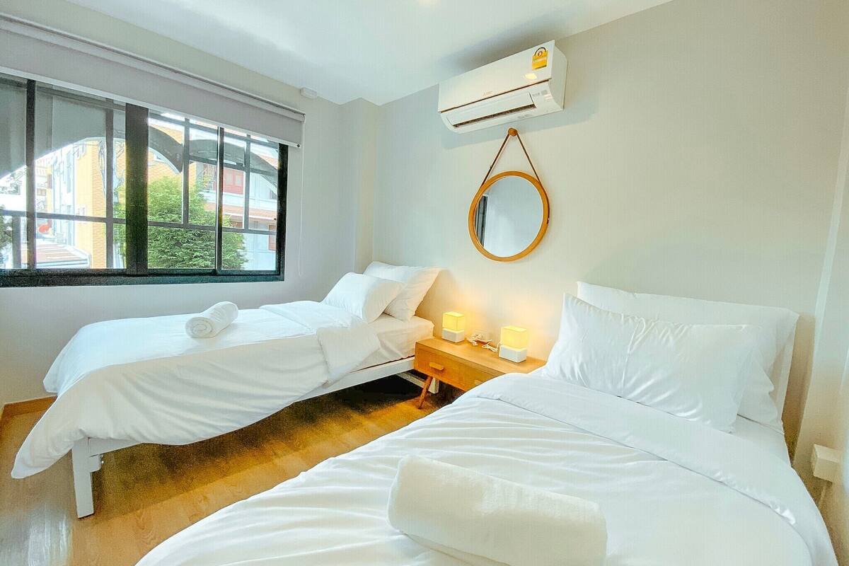 A hotel room with two single beds, a bedside table with lamps, a mirror, and an air conditioning unit.